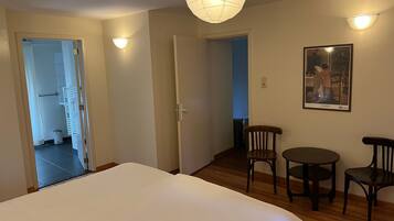 Habitación de hotel con una cama, una mesita, dos sillas, una lámpara de pared y una puerta que da a otra habitación.