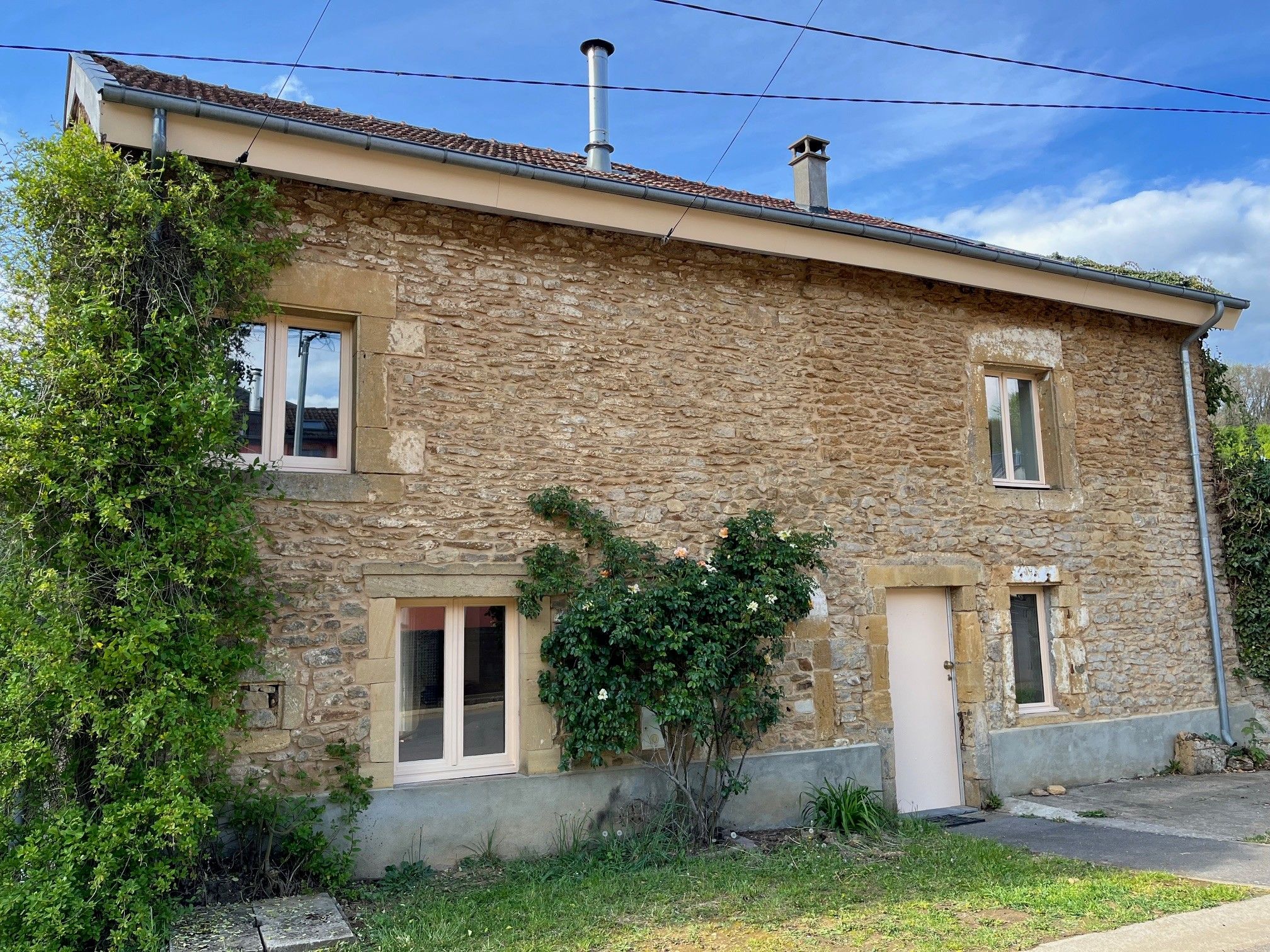 Une maison en pierre à deux étages, avec un toit en tuiles, une cheminée et un petit jardin agrémenté de verdure et d’un arbre.
