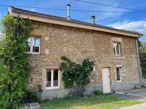 A two-story stone house with a tiled roof, a chimney, and a small garden area with greenery and a tree.