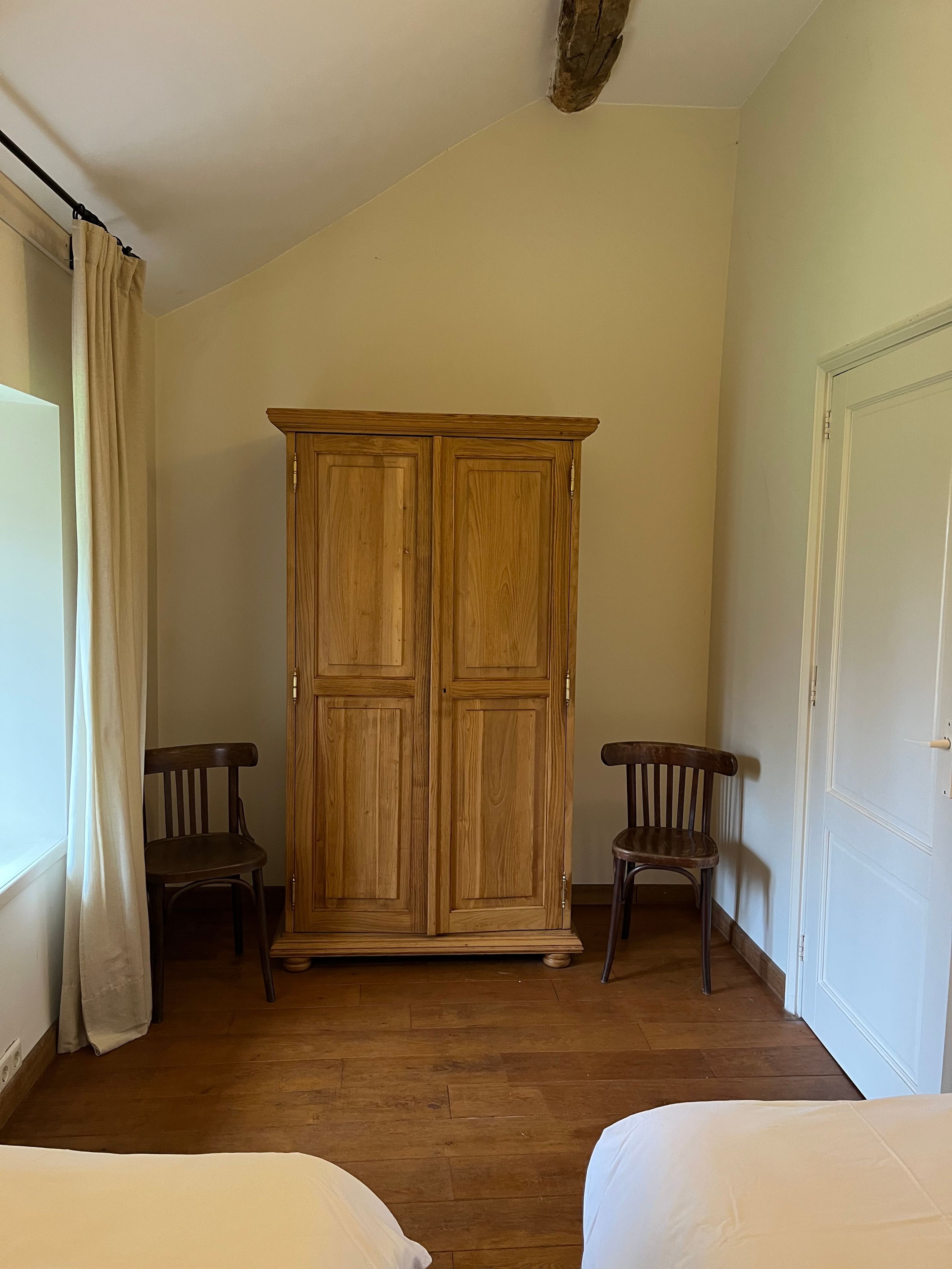 A room with a wooden wardrobe, two wooden chairs, a bed, and a white door.