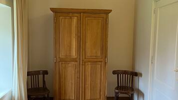 A room with a wooden wardrobe, two wooden chairs, a bed, and a white door.