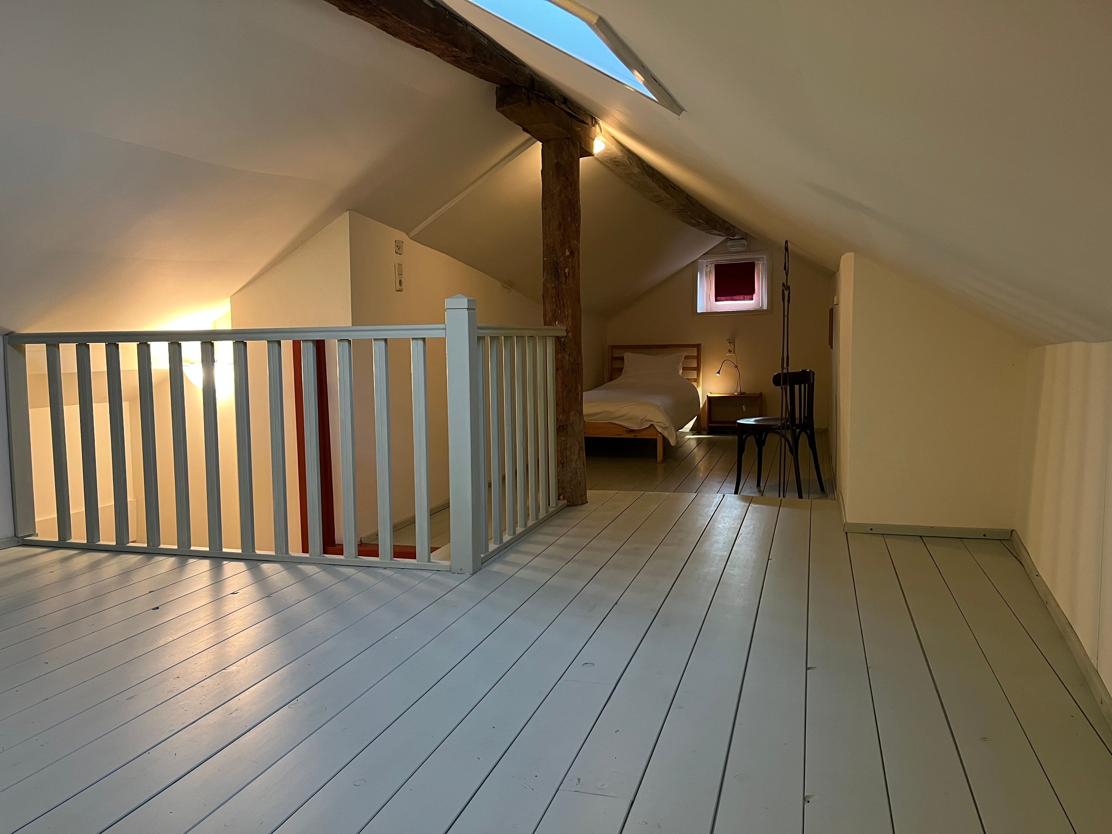 Une pièce avec un plafond en pente, un lit, une chaise, une table de chevet et une petite fenêtre.