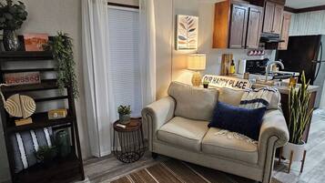 A cozy living room with a sofa, a small side table, a decorative shelf, and a kitchen area in the background.