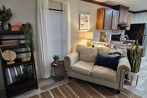 A cozy living room with a sofa, a small side table, a decorative shelf, and a kitchen area in the background.