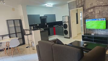 Una sala moderna con un sofá, un comedor con sillas, una cocina equipada y un televisor mostrando un partido deportivo.