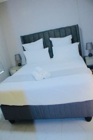Una cama bien tendida con sábanas blancas y almohadas, un cabecero oscuro y una lámpara de noche en una mesita.