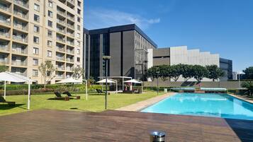A hotel pool area with a wooden deck, a modern building, and a high-rise apartment building in the background.