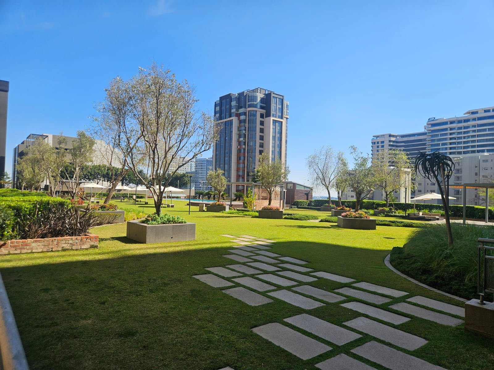 Un patio ajardinado con un sendero central, rodeado de vegetación y edificios modernos al fondo.