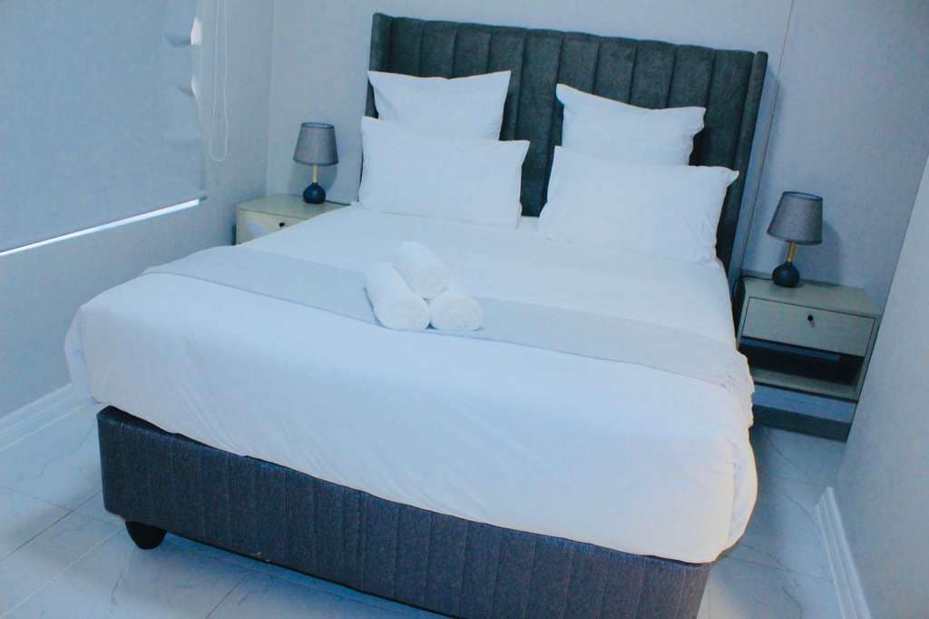 Una cama bien hecha con sábanas y almohadas blancas, dos mesitas de noche con lámparas y un par de pantuflas sobre la cama.