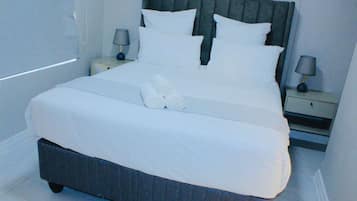 Una cama bien hecha con sábanas y almohadas blancas, dos mesitas de noche con lámparas y un par de pantuflas sobre la cama.