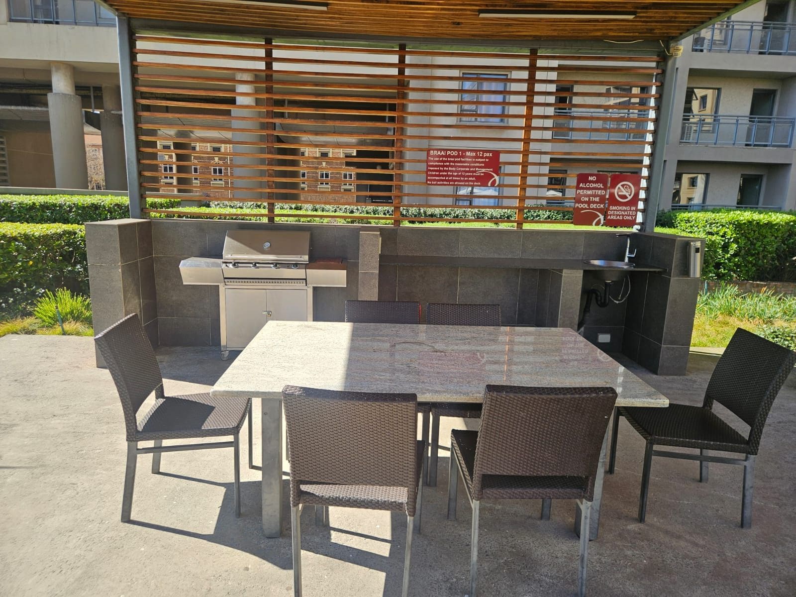 An outdoor barbecue area with a grill, seating, and a covered space with a view of the building behind.