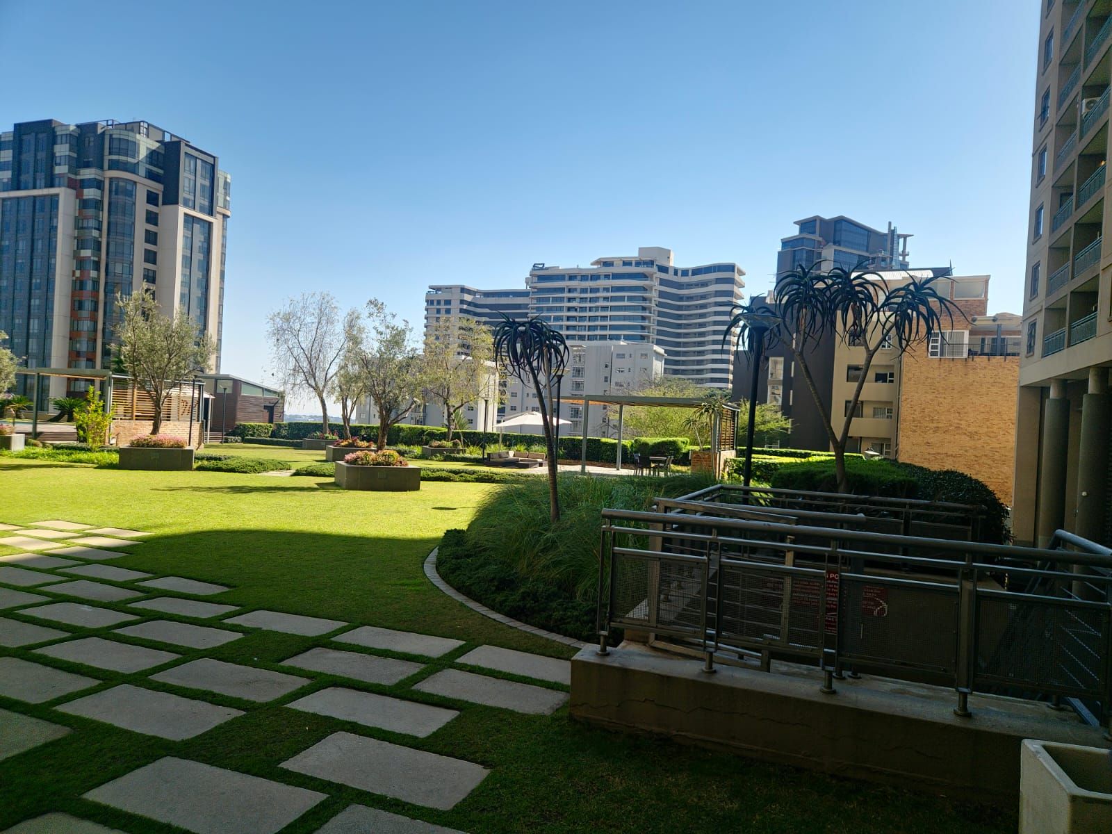Un patio con un sendero pavimentado, vegetación y edificios modernos al fondo.