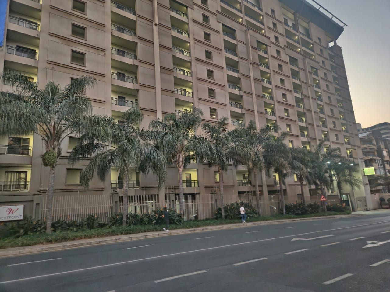 Un edificio residencial de varios pisos con balcones, una entrada vigilada y una calle en frente.