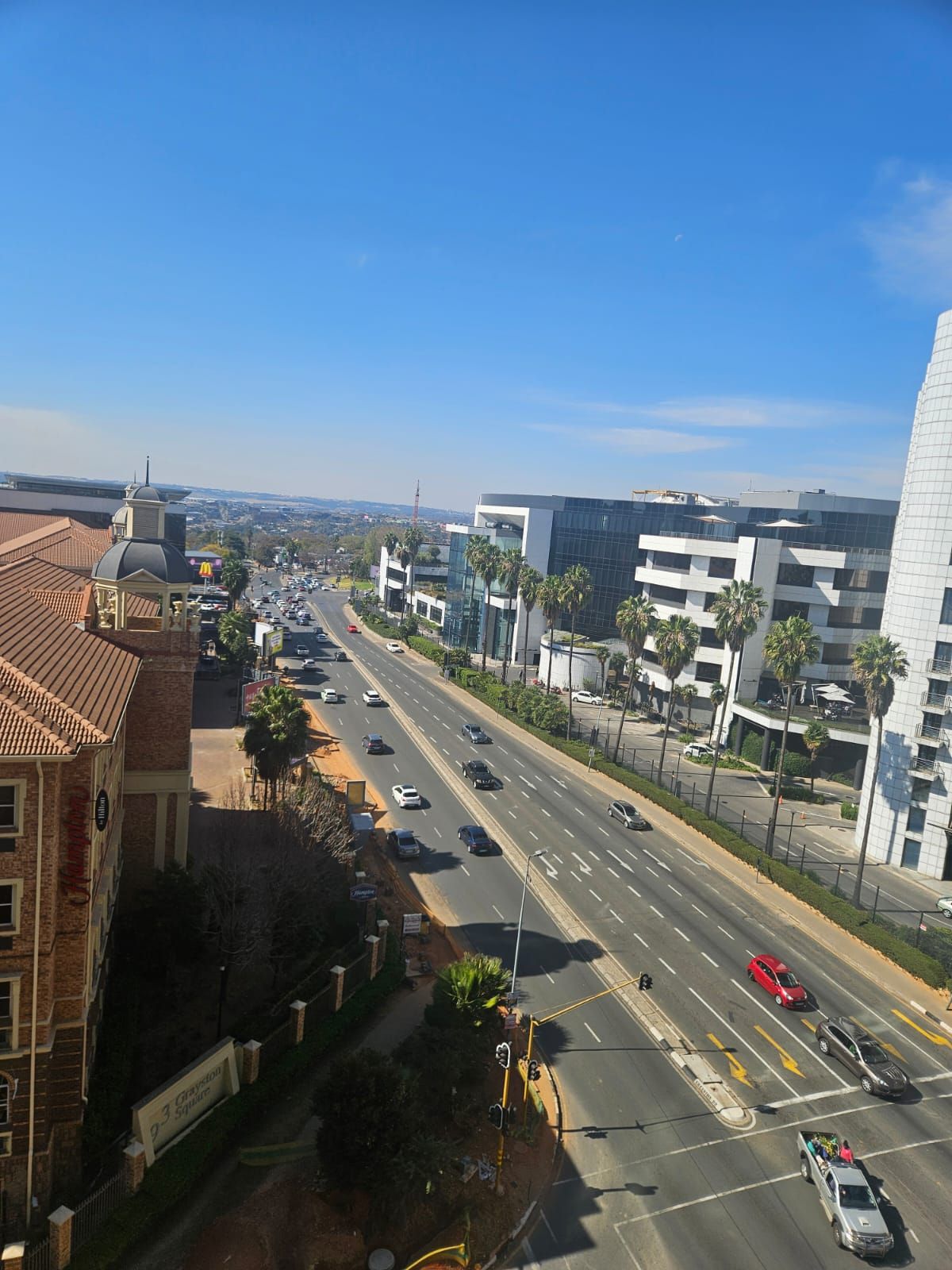 A multi-lane road with modern buildings, a clear sky, and a distant view of a cityscape.