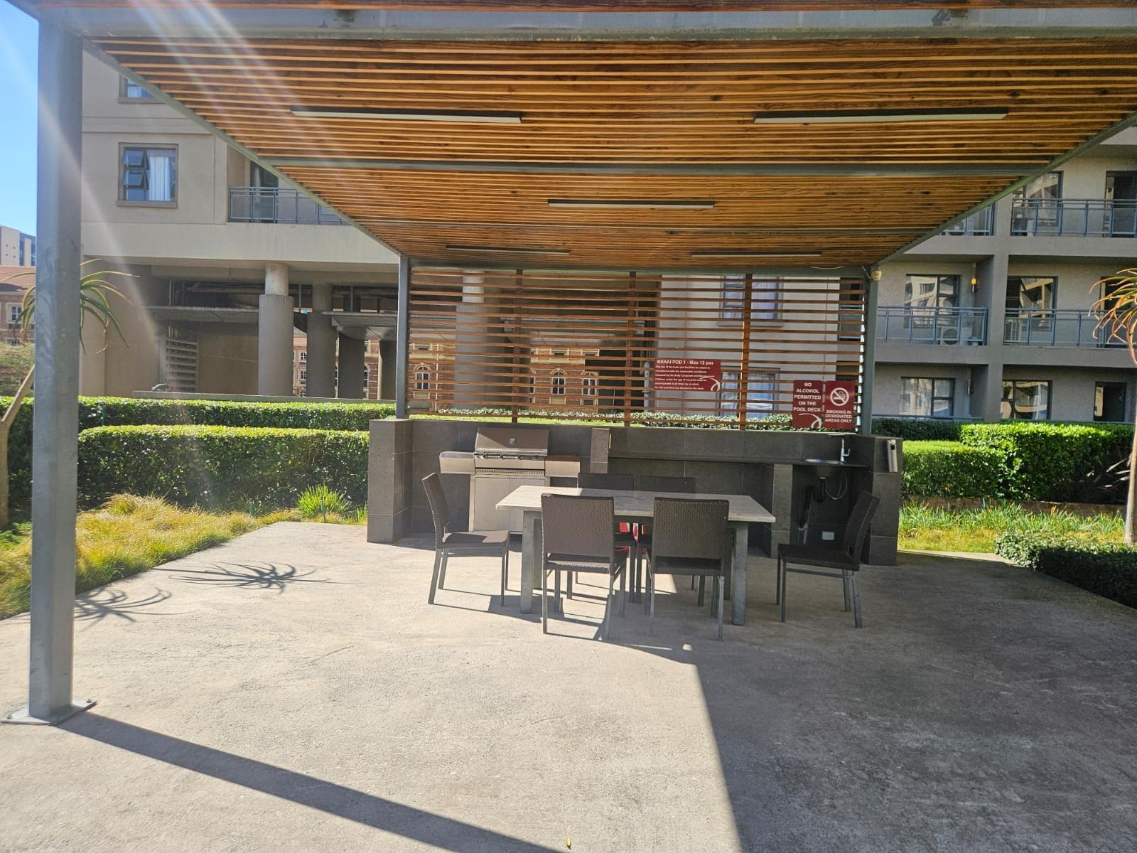 An outdoor seating area with tables and chairs, a wooden pergola overhead, and a pool deck in the background.