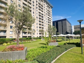 A modern hotel with a large building, a courtyard with greenery, and outdoor seating. - Sandton Westpoint Luxury Apartments - 7 (Sandton)