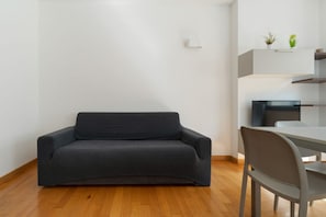 A modern living room with a dark-colored sofa, a dining table with chairs, and a white wall with a small shelf holding two potted plants. - GuestHost - Cozy Central Apartment in Milan (Milan)