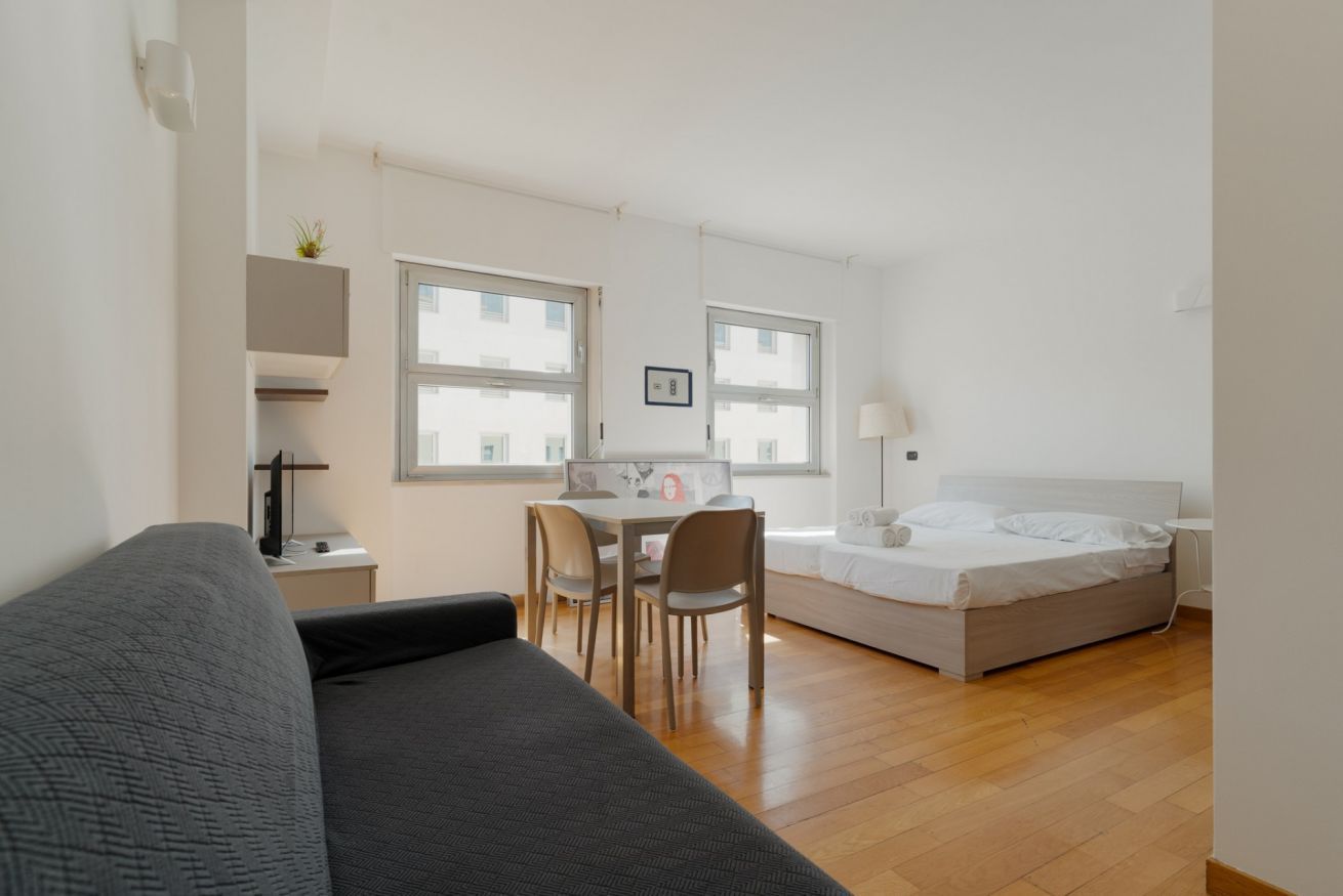 Habitación de hotel compacta con una cama, un escritorio con sillas, un sofá y ventanas amplias.