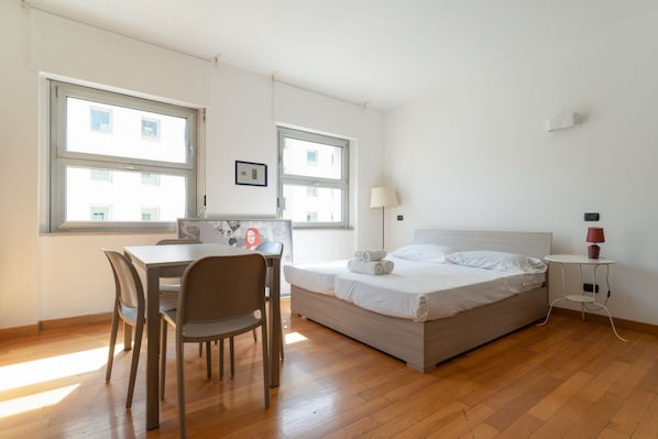 Una habitación con cama, escritorio y silla. Dos ventanas dejan entrar luz natural.