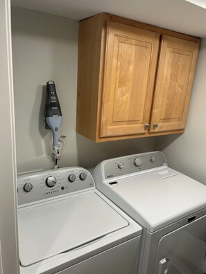 A laundry area with two white washing machines and a dryer, a wooden cabinet above, and a hairdryer on the wall. - Ocean View, Golf Cart Included, Pet Friendly, Walk to the Beach - 406 Captain John Fripp Villa (Fripp Island)