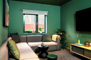 A living room with a green wall, a grey sofa, a window with plants, and a TV mounted on the wall. - Student Only Station Court (London)