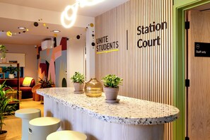 A reception area with a sign reading 'Station Court' and a 'Staff only' door. There are potted plants on the counter and a seating area with a red chair. - Student Only Station Court (London)