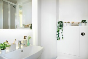 A bathroom with a white sink, a mirror, a shower, and a toilet. There are plants and toiletries on the wall. - Student Only Station Court (London)