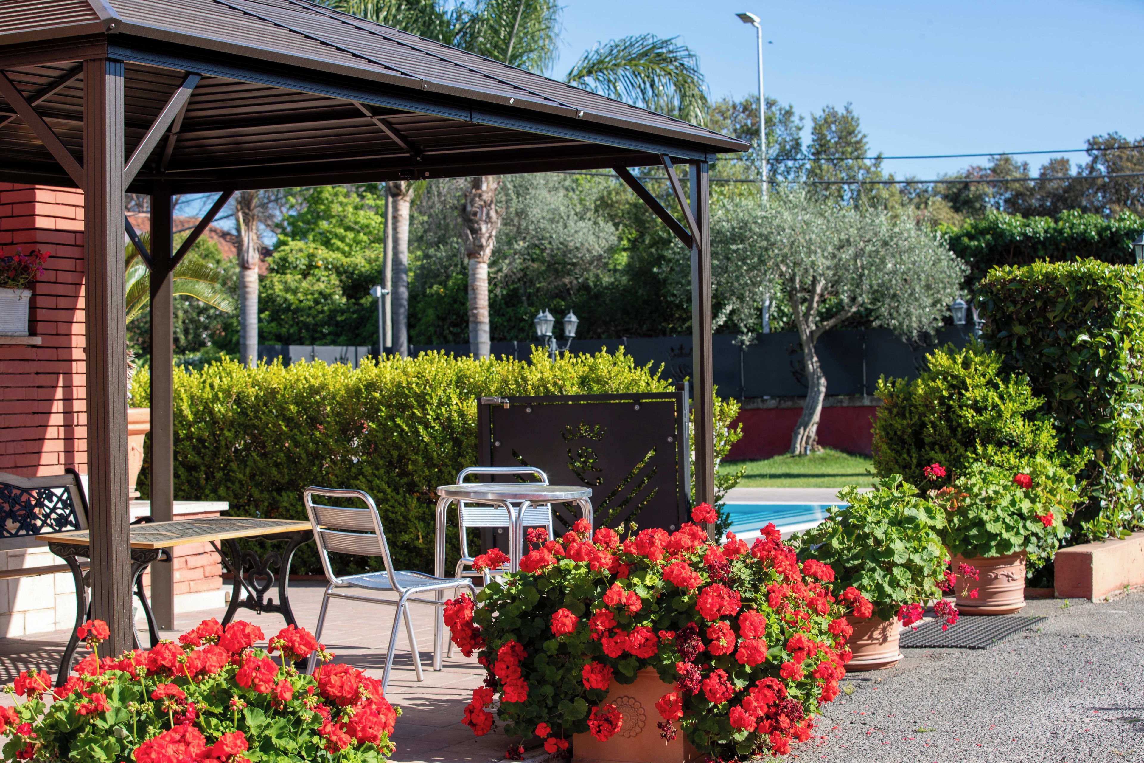 Un espace extérieur aménagé, abrité et doté de chaises en métal et d’une table, entouré de plantes en pot et d’une piscine.