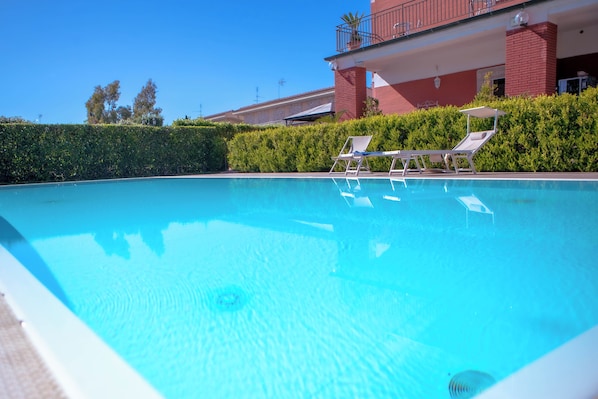A clear swimming pool with lounge chairs and a hedge in the background.