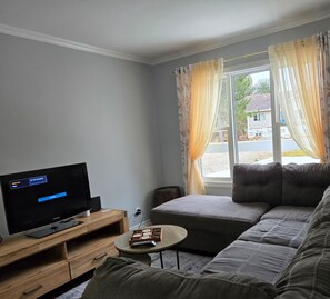 Un salon avec un téléviseur à écran plat posé sur un meuble en bois, un canapé gris, une petite table ronde sur laquelle se trouve un jeu de société, et une fenêtre avec des rideaux.