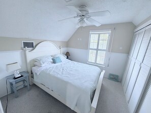 Quarto com cama, mesa de cabeceira, abajur, ventoinha no teto, janela com persianas e radiador.
