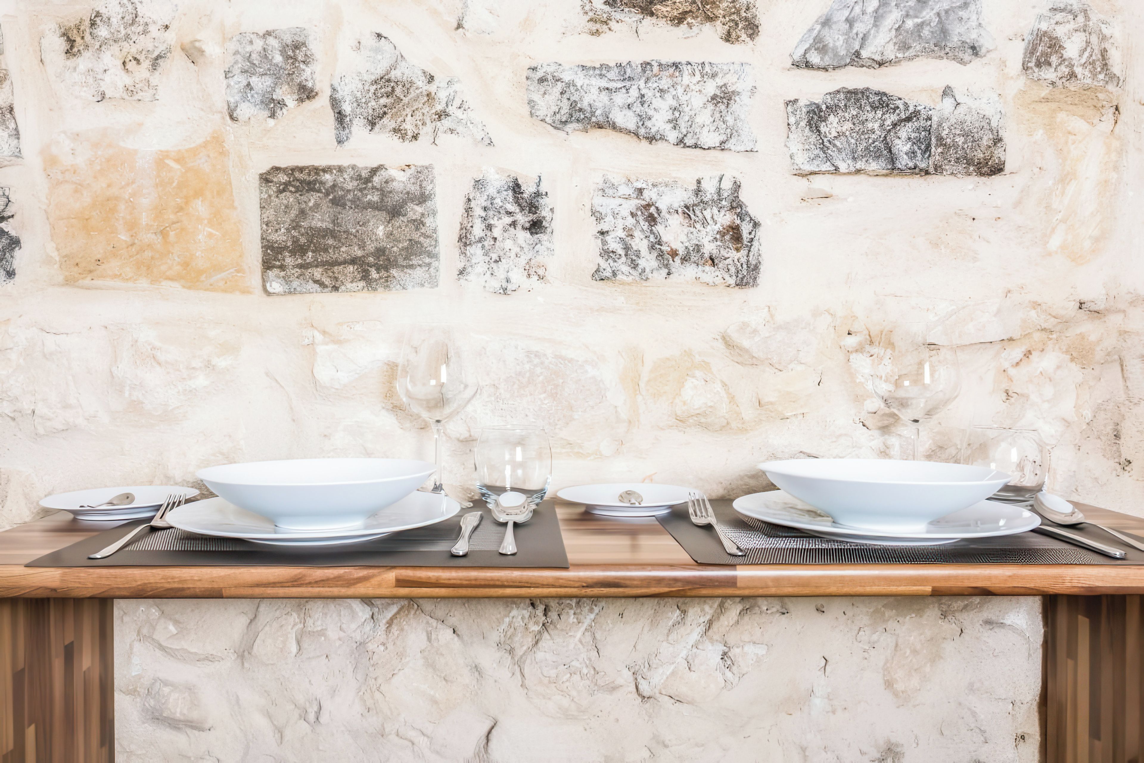 A dining table set with white plates, bowls, and cutlery against a stone wall.