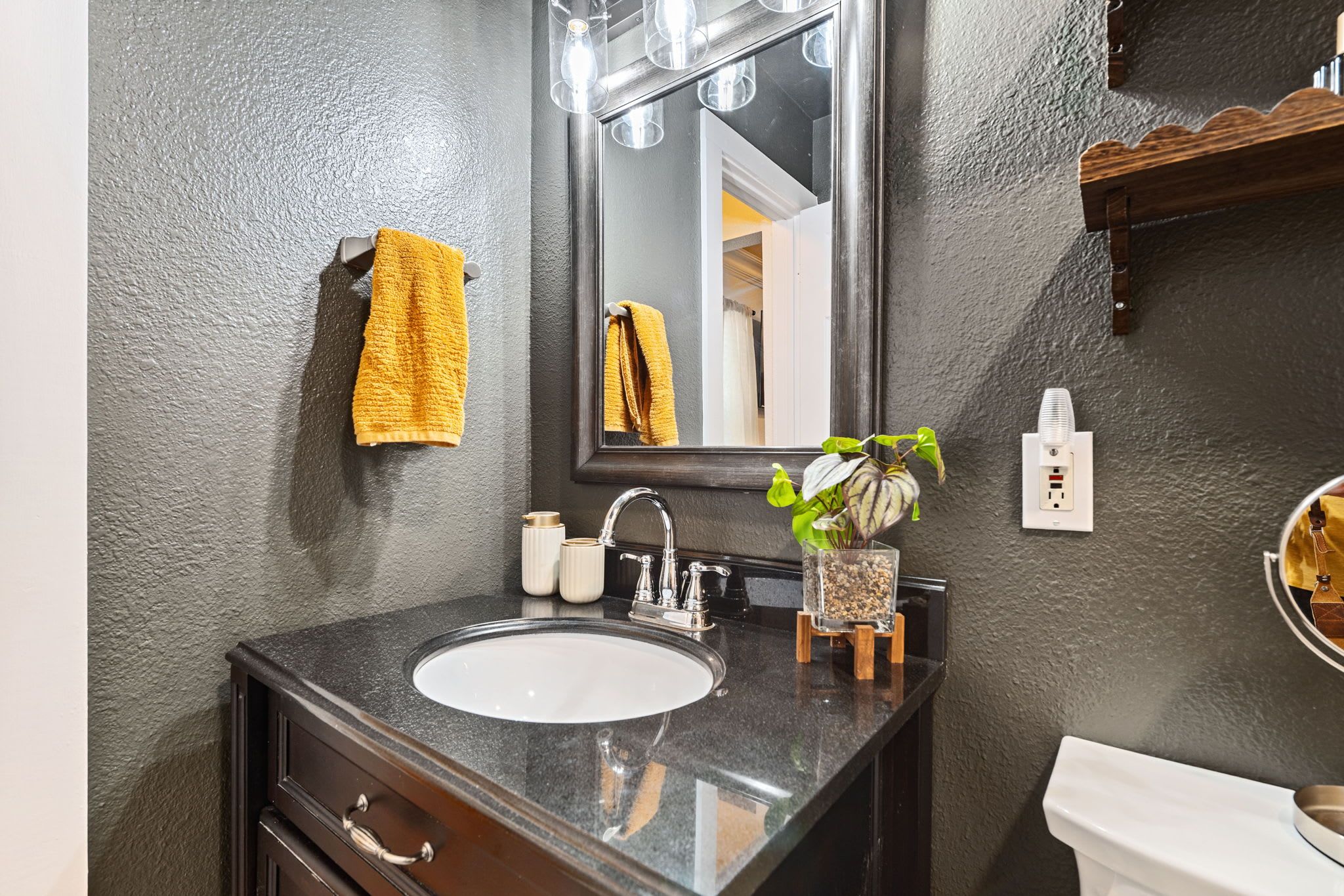 Une salle de bain avec un lavabo en marbre foncé, un miroir, une serviette jaune, une petite plante et des toilettes blanches.