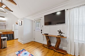 A modern living room with a wooden floor, a flat-screen TV mounted on the wall, a rustic wooden coffee table, and a kitchen area with a refrigerator and sink.