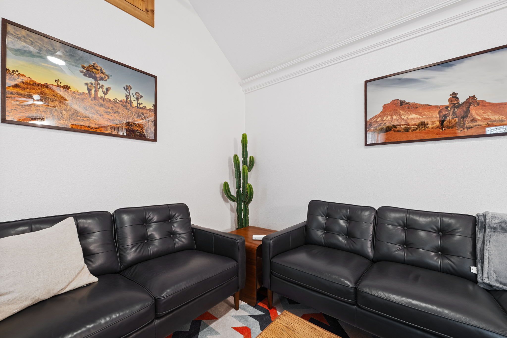 A living room with two black leather sofas, a small wooden side table, a cactus plant, and two framed desert landscape paintings on the walls.