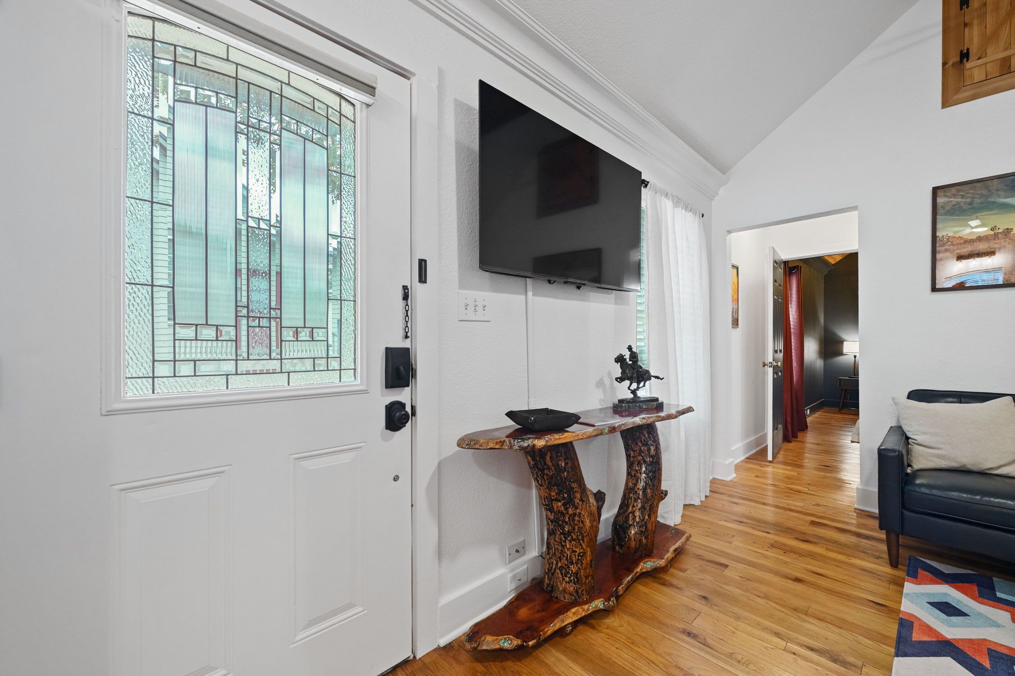 Un pasillo con una puerta blanca, un televisor de pantalla plana montado, un mueble consola de madera con una escultura y una alfombra estampada.