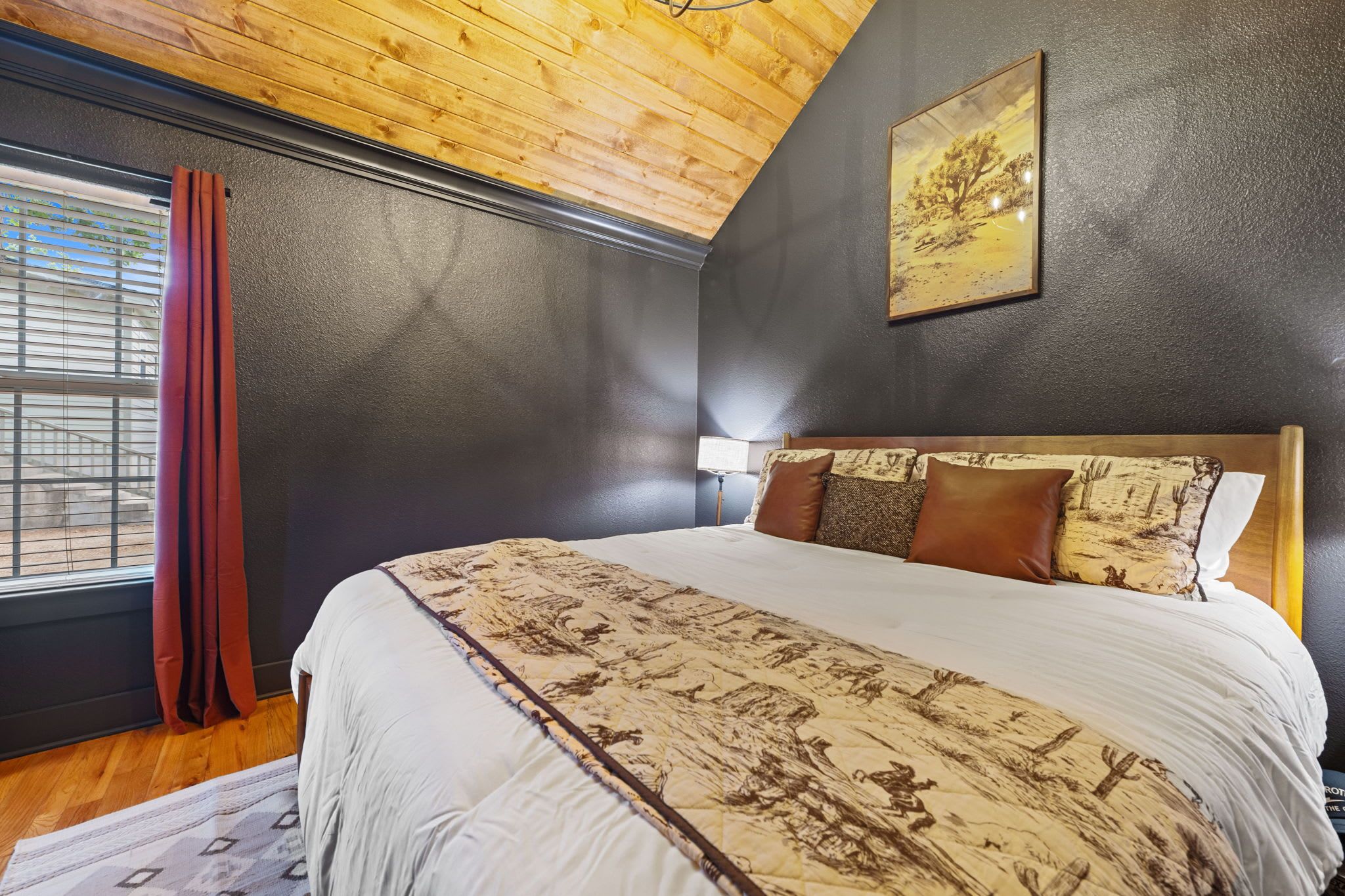Een slaapkamer met een groot bed, een raam met gordijnen, een hoofdbord met kussens en een schilderij aan de muur.