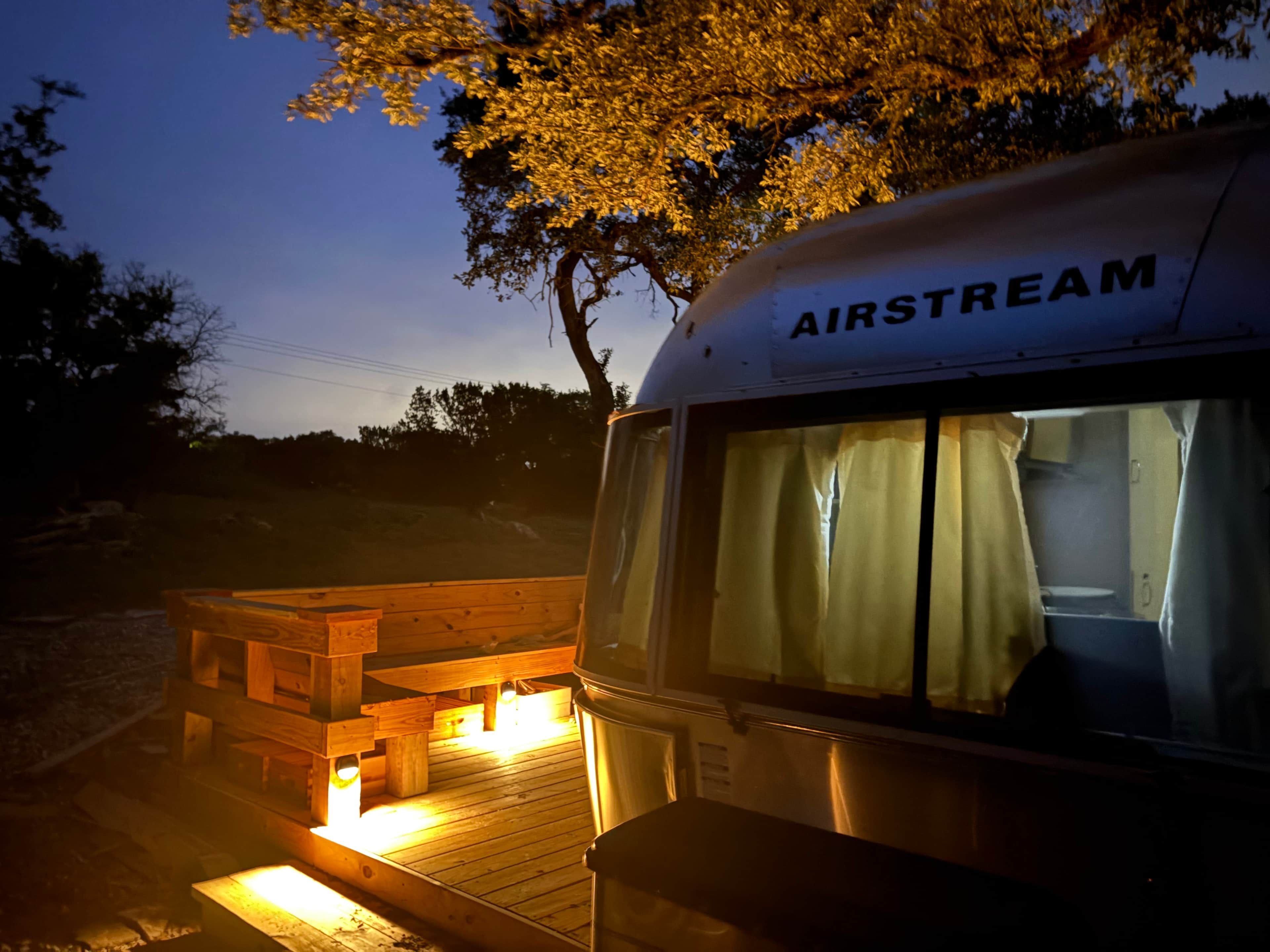A stationary RV with the name 'AIRSTREAM' on its side, a wooden deck, and a window with curtains.