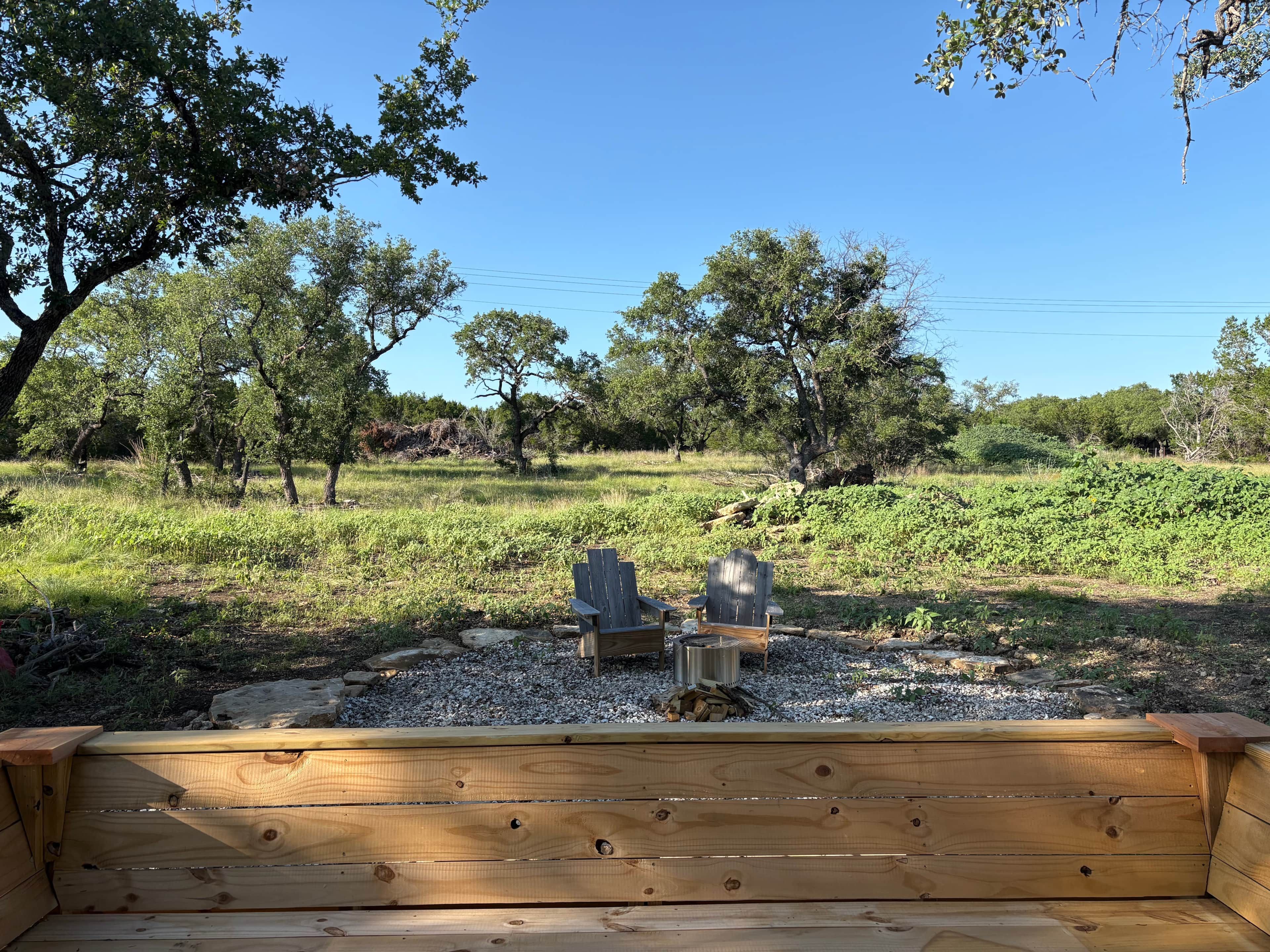 Un banc en bois, accompagné de deux fauteuils Adirondack, est placé face à un paysage naturel composé d’arbres et d’un ciel dégagé.