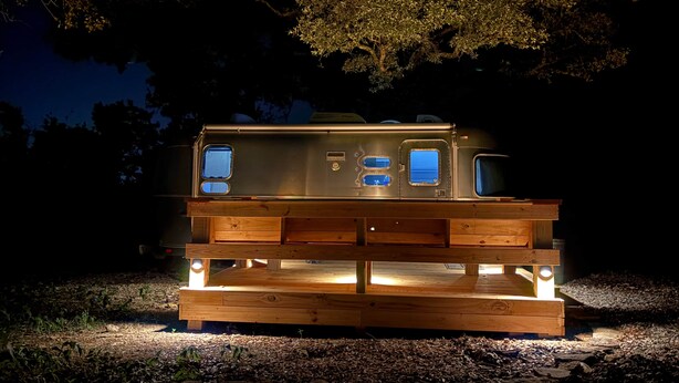 Una caravana estacionada con una terraza de madera y varias ventanas, rodeada de árboles.