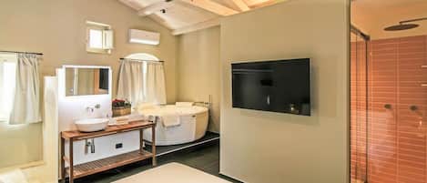 A modern hotel room with a large bed, a vanity with a mirror, a bathtub, and a flat-screen TV mounted on the wall.