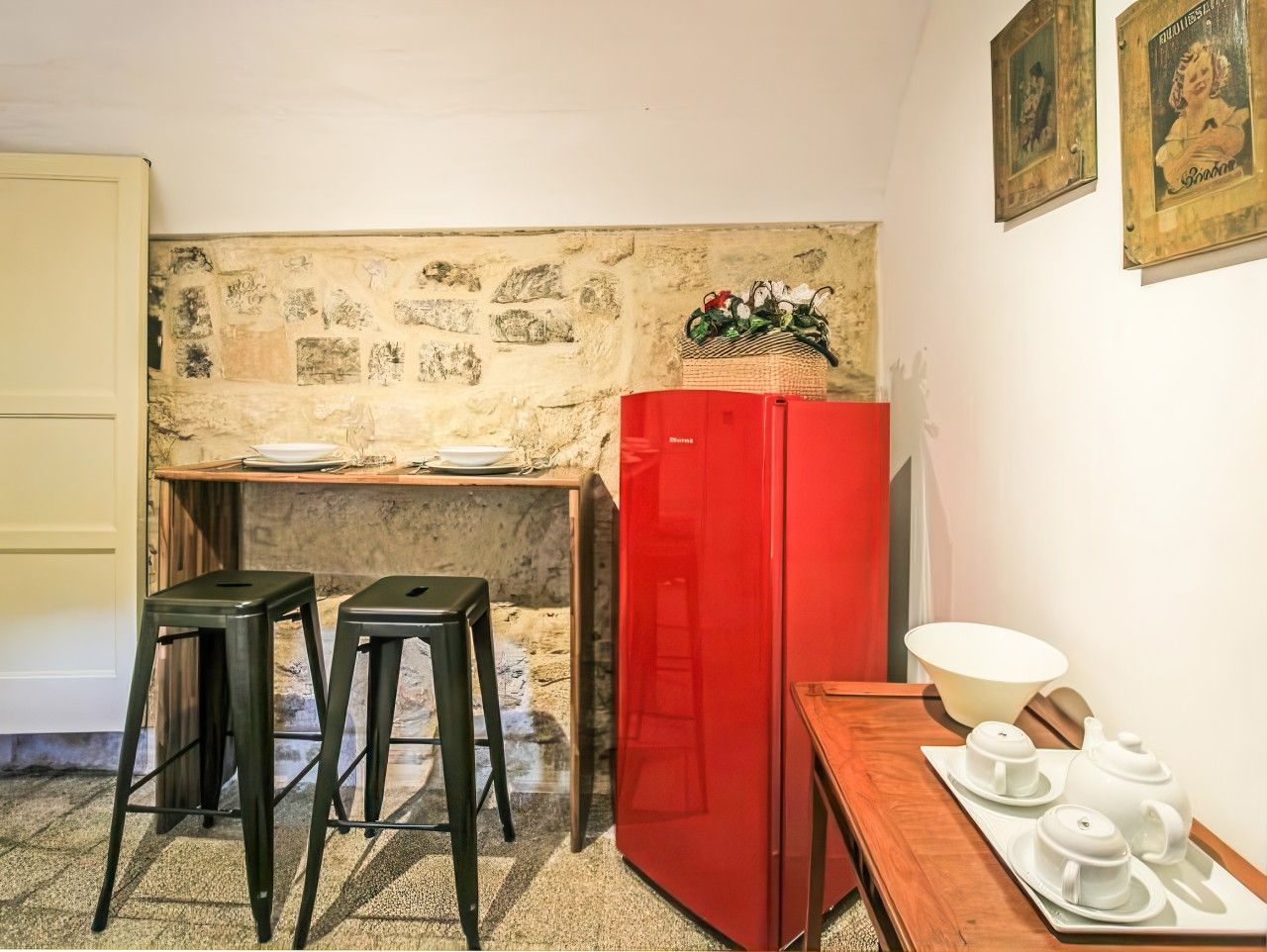 Une cuisine compacte avec un réfrigérateur rouge, deux tabourets, une table en bois avec un service à thé et un mur en pierre.