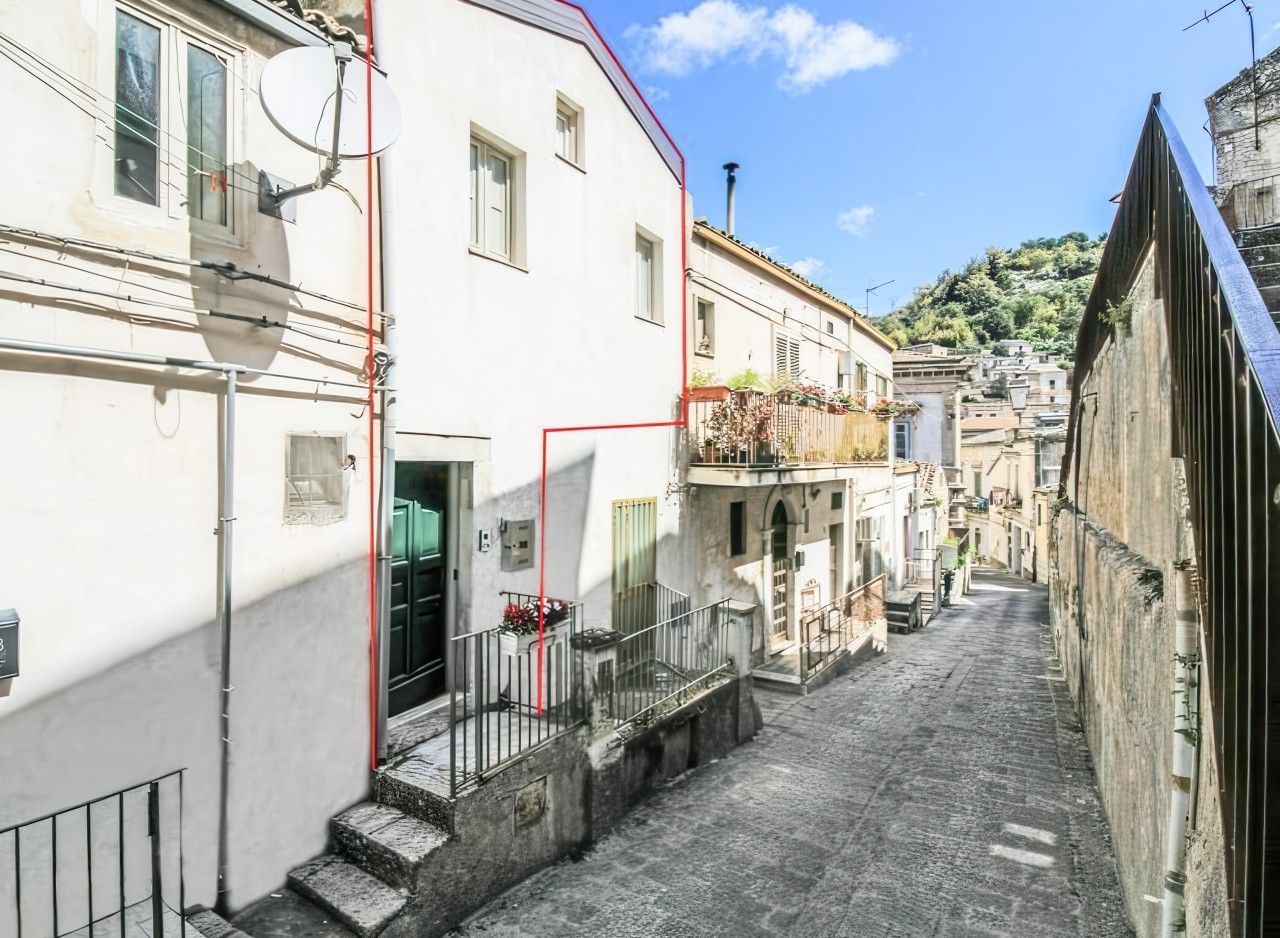 Une rue étroite bordée de bâtiments des deux côtés, un balcon bien visible orné de plantes et un ciel dégagé.