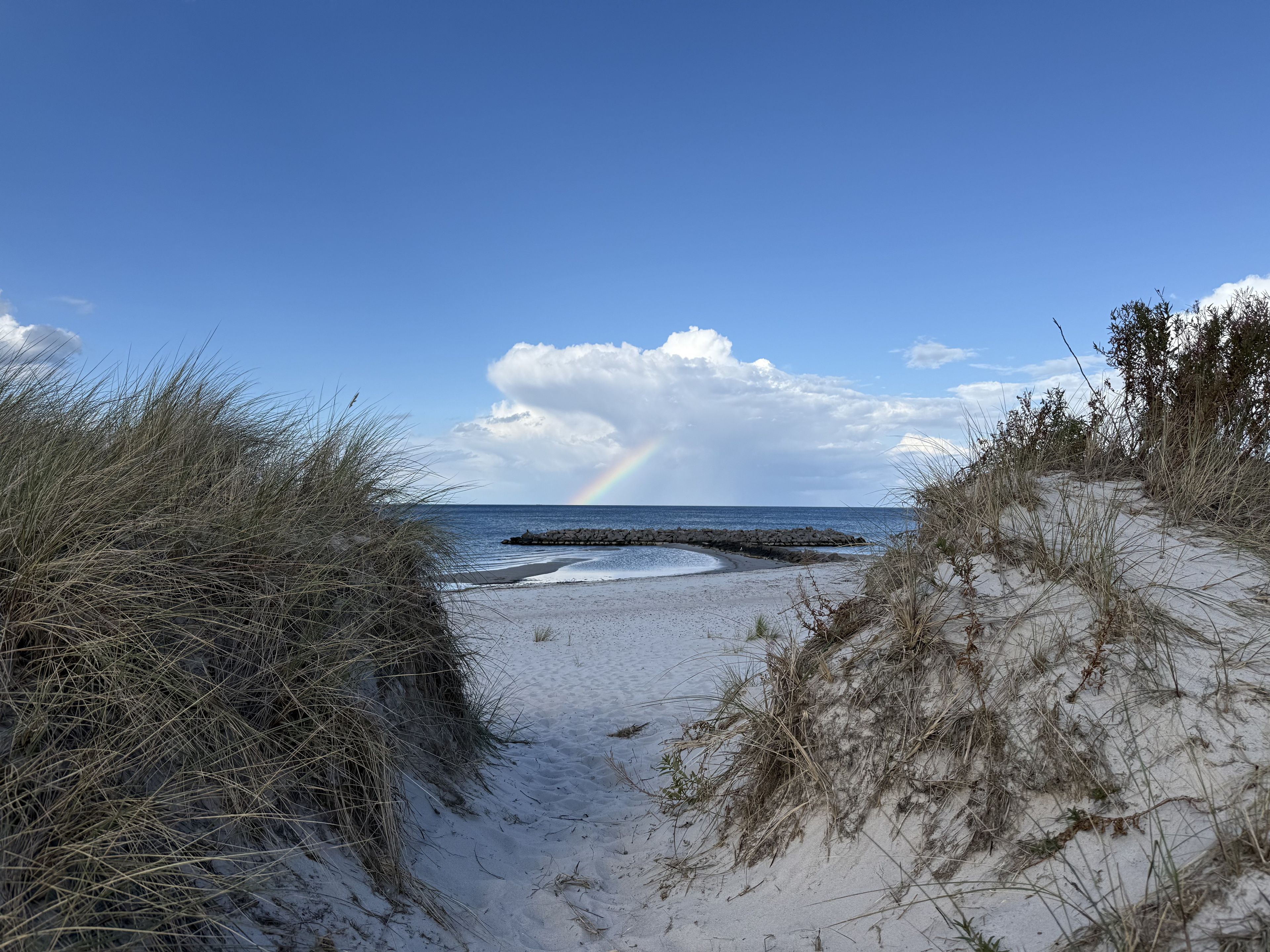 Ein Strandweg führt zum Meer, in der Ferne ist ein Regenbogen zu sehen.