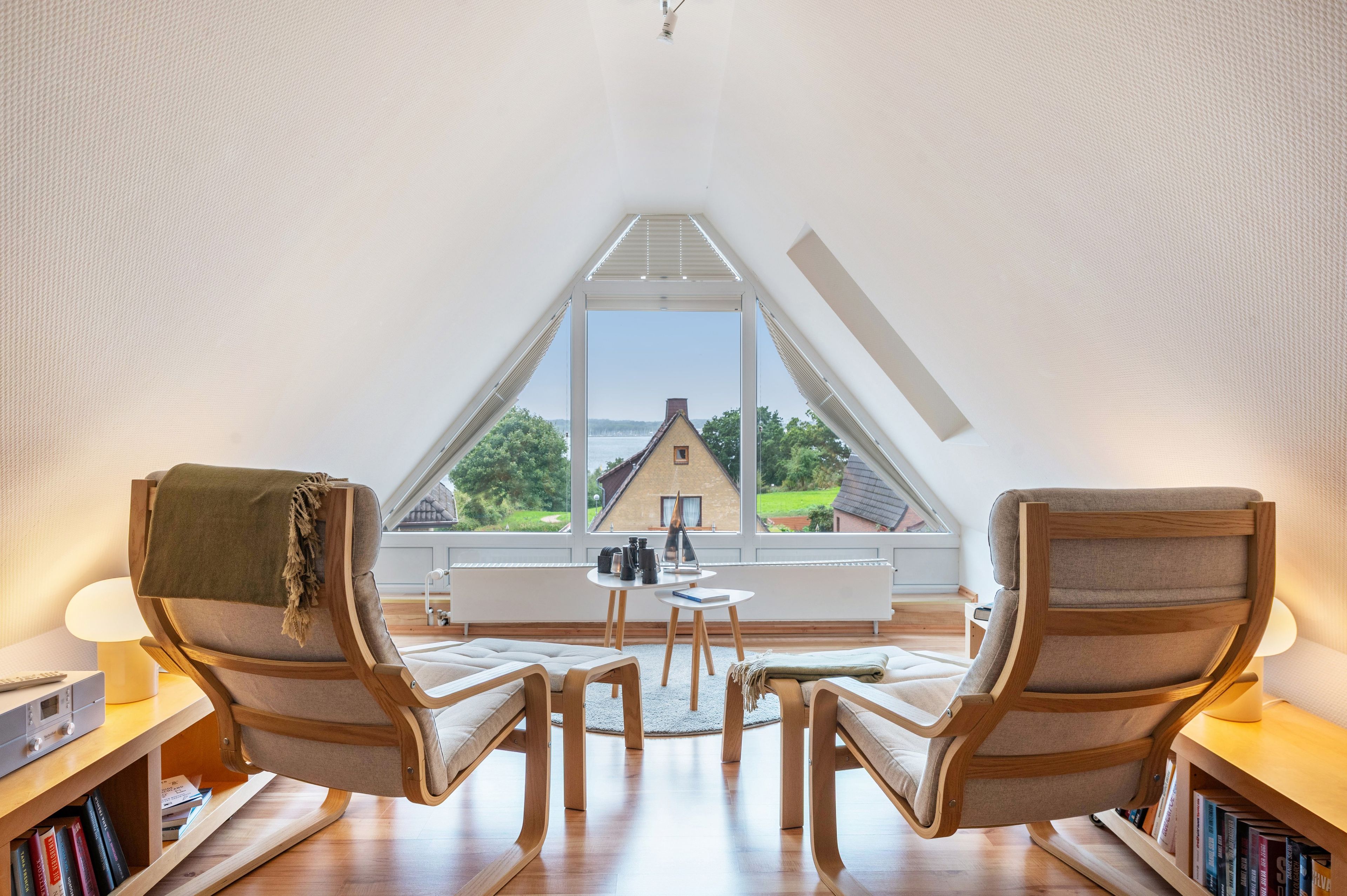 Quarto com teto inclinado, duas cadeiras de madeira, uma mesa pequena e vista para uma casa através de janelas amplas.