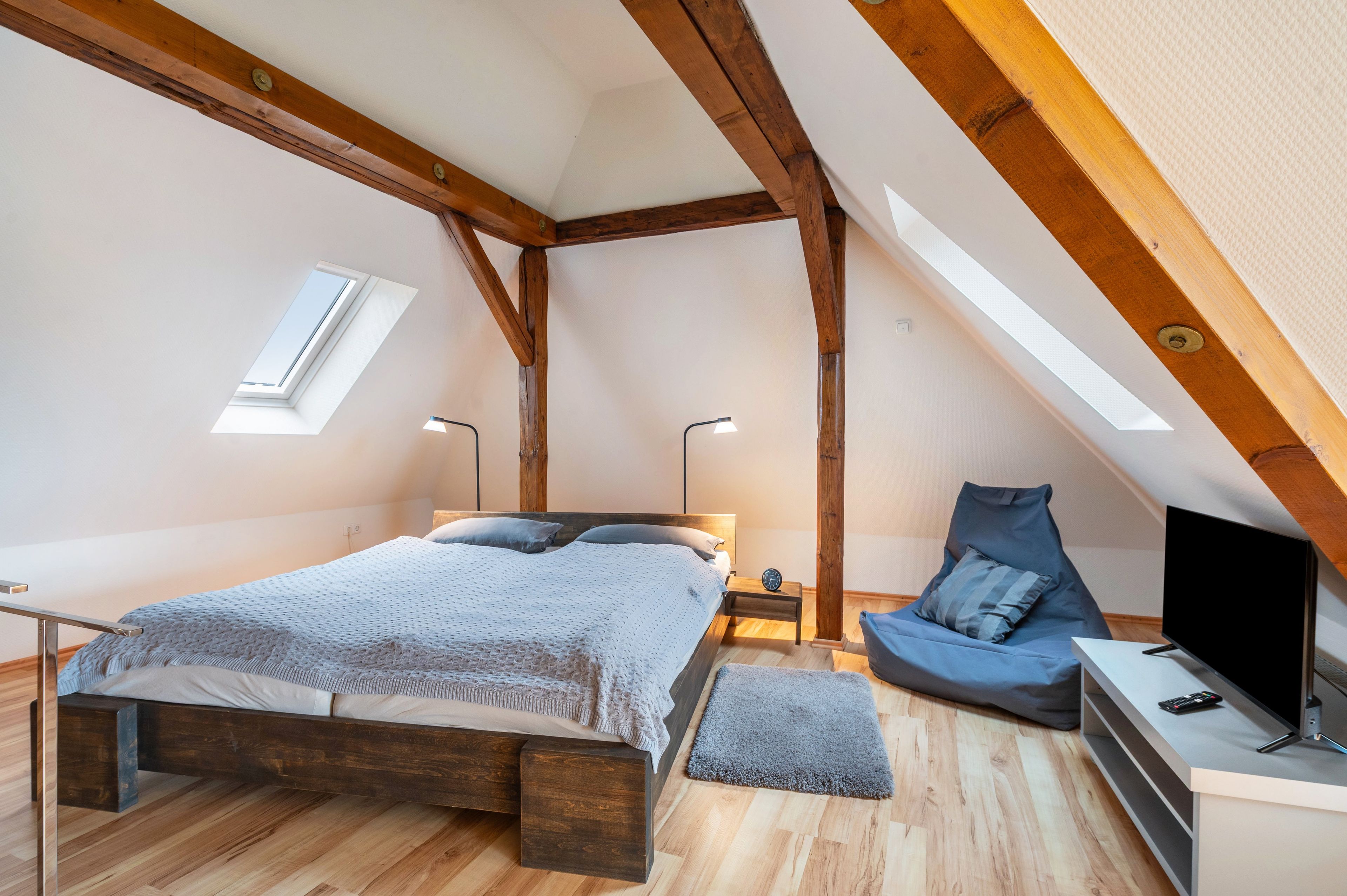 Ein Schlafzimmer mit schrägen Wänden, Holzbalken, einem Bett mit grauer Decke, einem Nachttisch mit Lampe, einem blauen Sessel und einem Fernseher auf einem Schrank.
