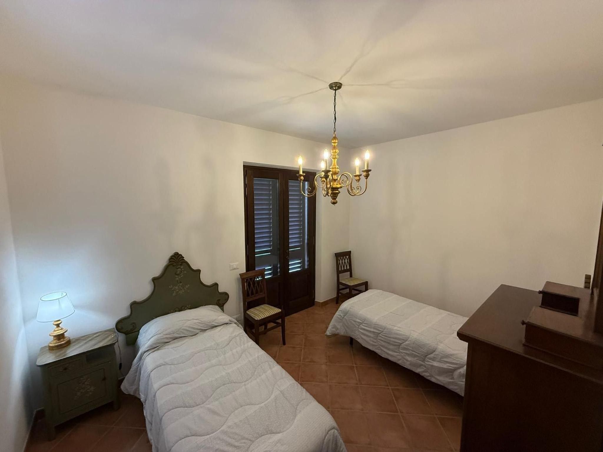 Quarto com duas camas de solteiro, mesa de cabeceira com abajur, cadeira de madeira e cômoda.