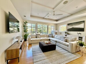 Ein modernes Wohnzimmer mit einer Eckcouch, einem Flachbildfernseher, einem Deckenventilator und großen Fenstern.