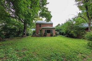 A brick house with a small porch and a covered area, surrounded by a grassy yard and trees. - Charming Shepherdstown Home - Walk to Downtown (Shepherdstown)