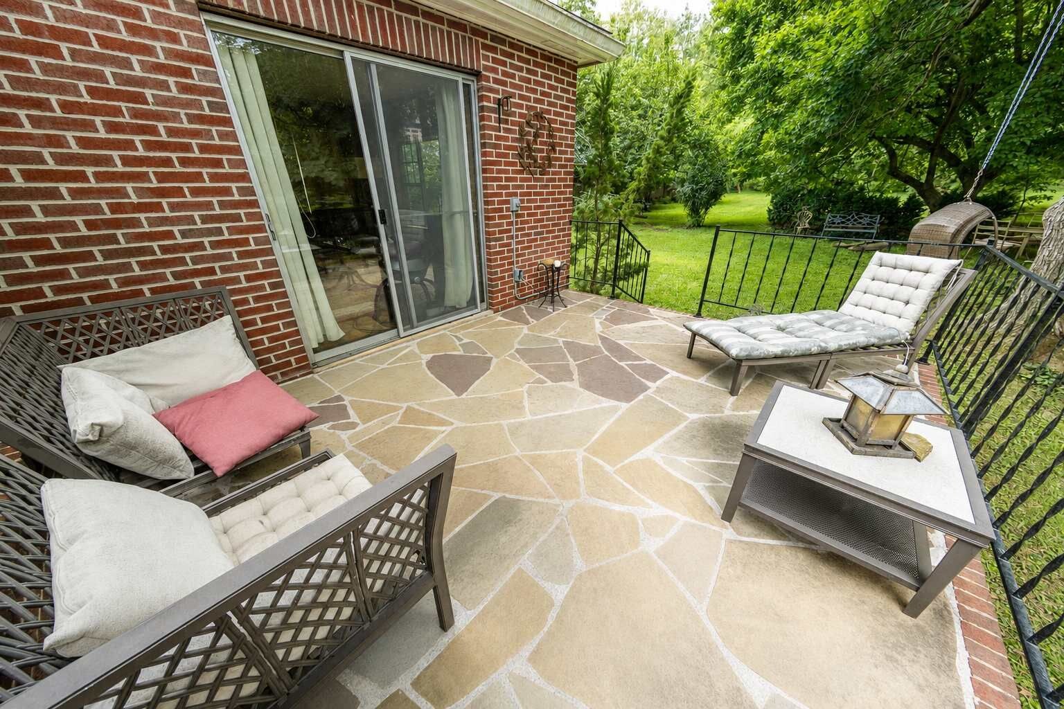An outdoor patio with stone flooring, two cushioned chairs, a small table with a lantern, and a brick building with a sliding glass door.
